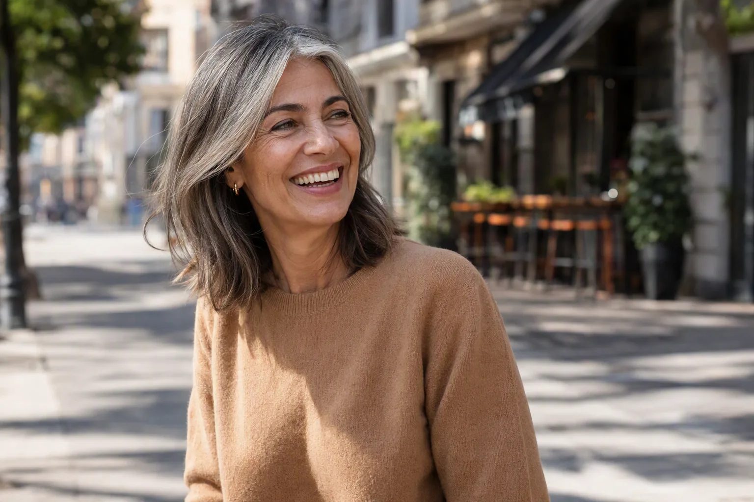Mujer de unos 55 años con melena midi en capas y canas naturales, sonriendo en una calle soleada