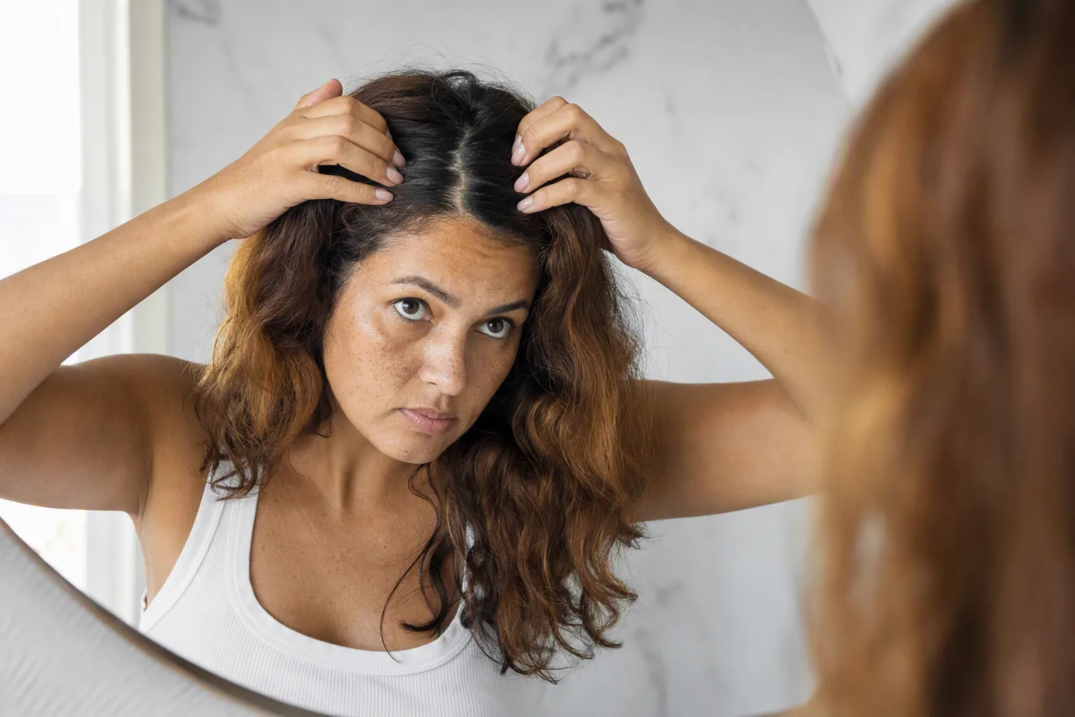 Mulher repartindo o cabelo diante do espelho para mostrar a raiz escura crescida sobre o comprimento tingido